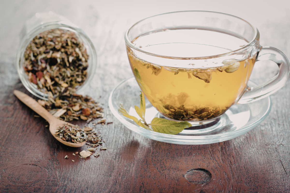 A clear glass cup of herbal tea on a saucer sits on a wooden table next to a jar of loose tea leaves and a wooden spoon with tea leaves.