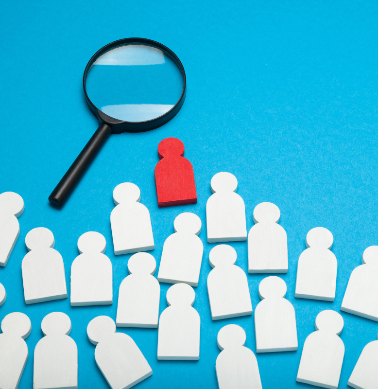 A red figure stands out among white figures, with a magnifying glass above it, symbolising focus and analysis in a crowd.