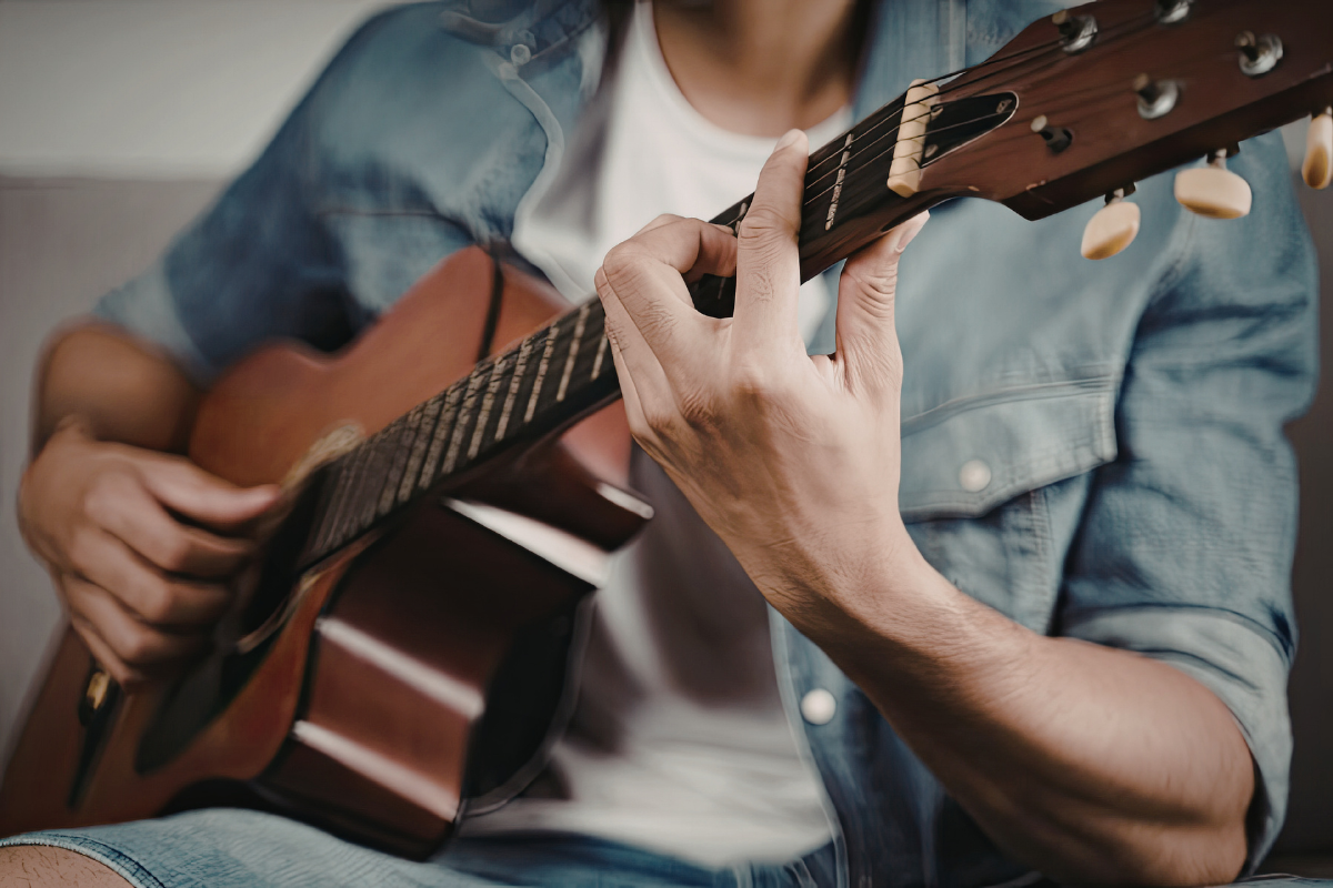 Person in a denim shirt sitting and playing an acoustic guitar, with focus on their hands and the guitar.