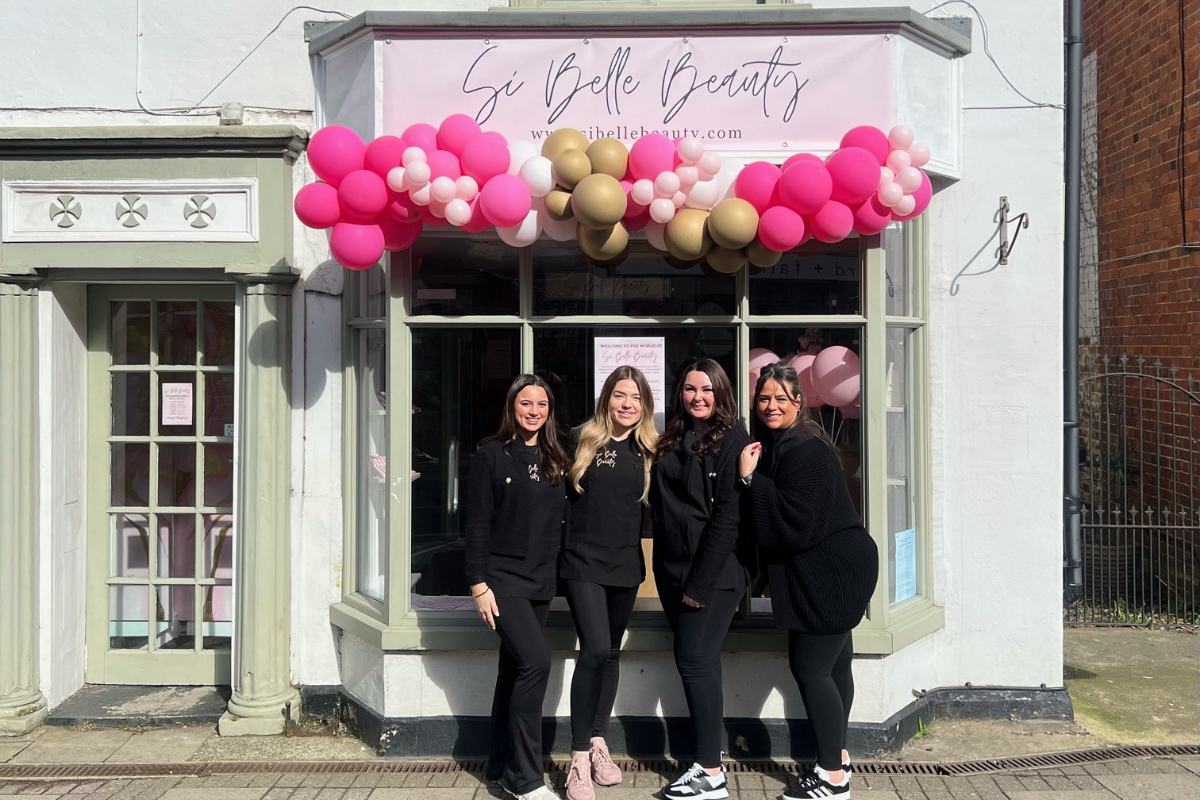 A beauty salon storefront adorned with pink and gold balloons, featuring four staff members in black outfits posing together.